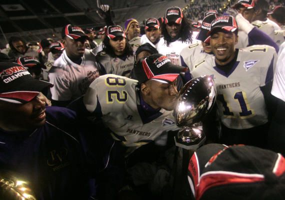 A jubilant group of football players in purple and white uniforms celebrate with a championship trophy amidst cheers and excitement.